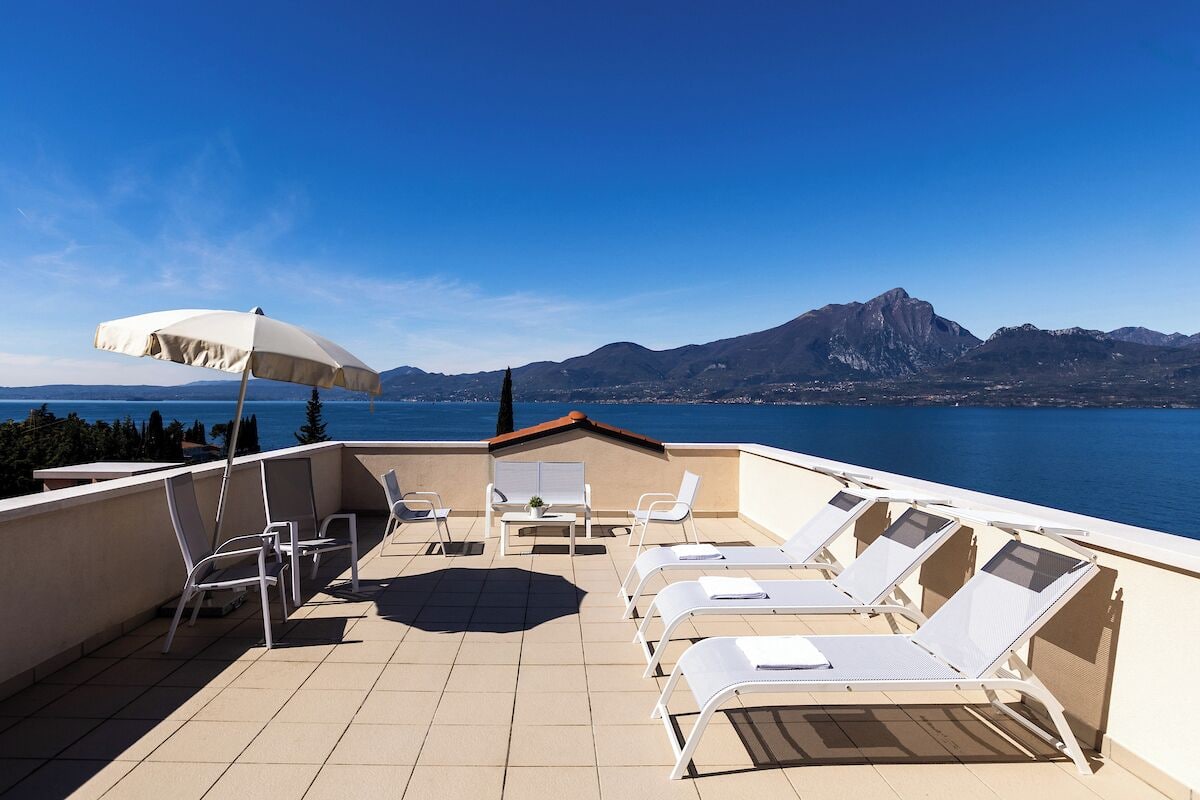 A terrace with sun loungers and a table, overlooking a body of water and mountains.