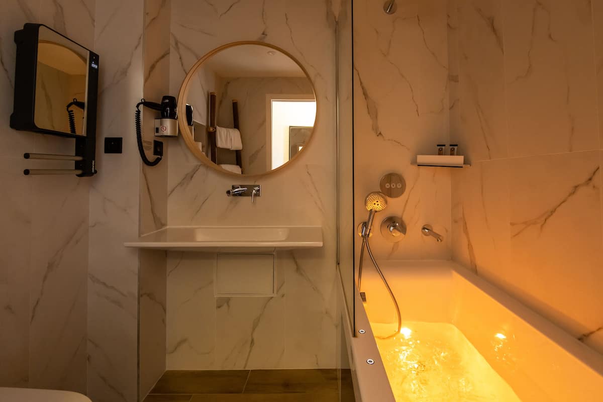 Un baño moderno con pared de mármol, un espejo redondo, lavabo empotrado y bañera con ducha de mano.