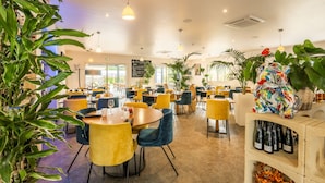 Een ruim restaurant met gele en blauwe stoelen, diverse planten en een wijnrek.