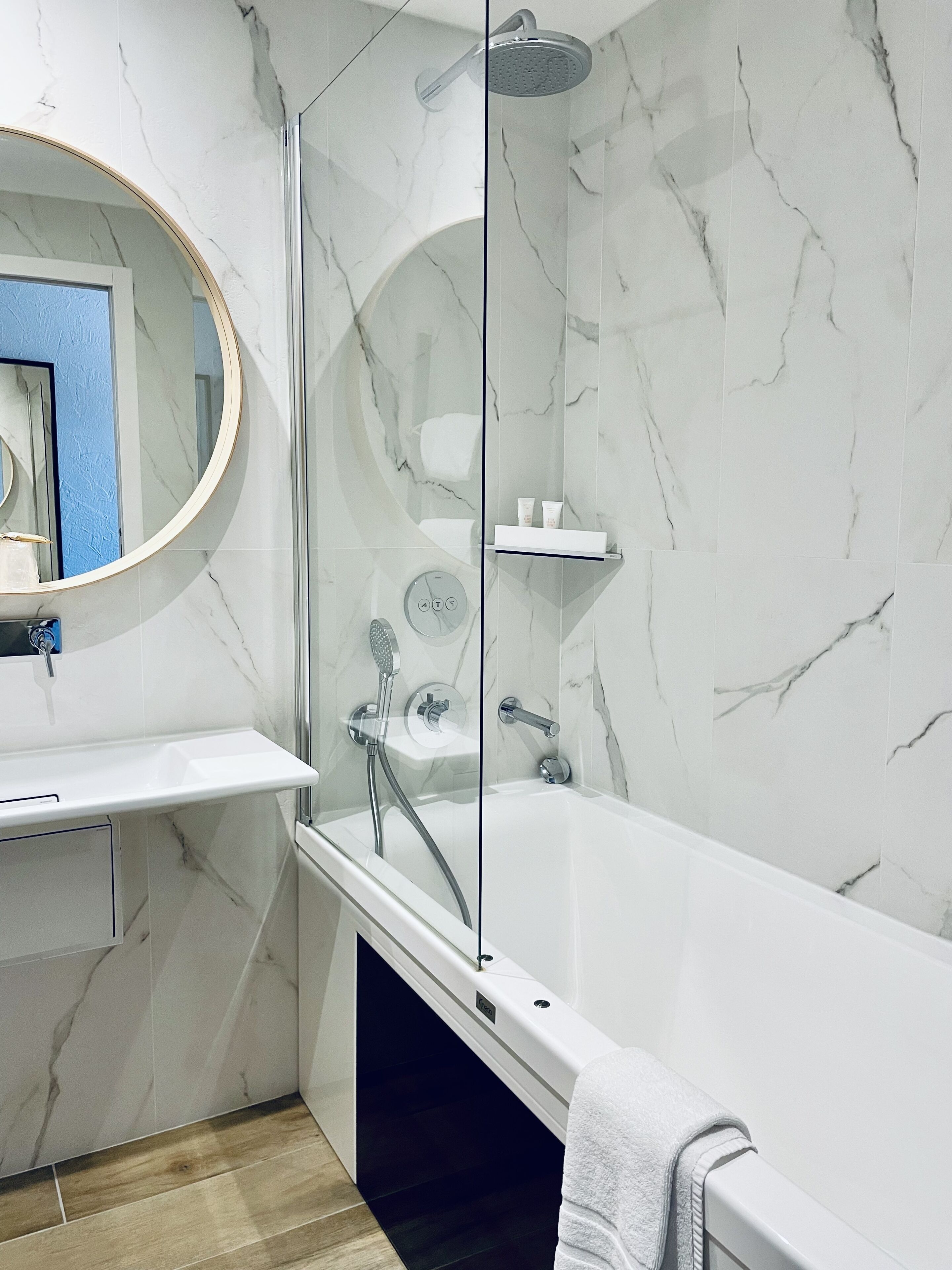 A modern bathroom with a marble wall, a round mirror, a white sink, and a bathtub with a glass-enclosed shower.