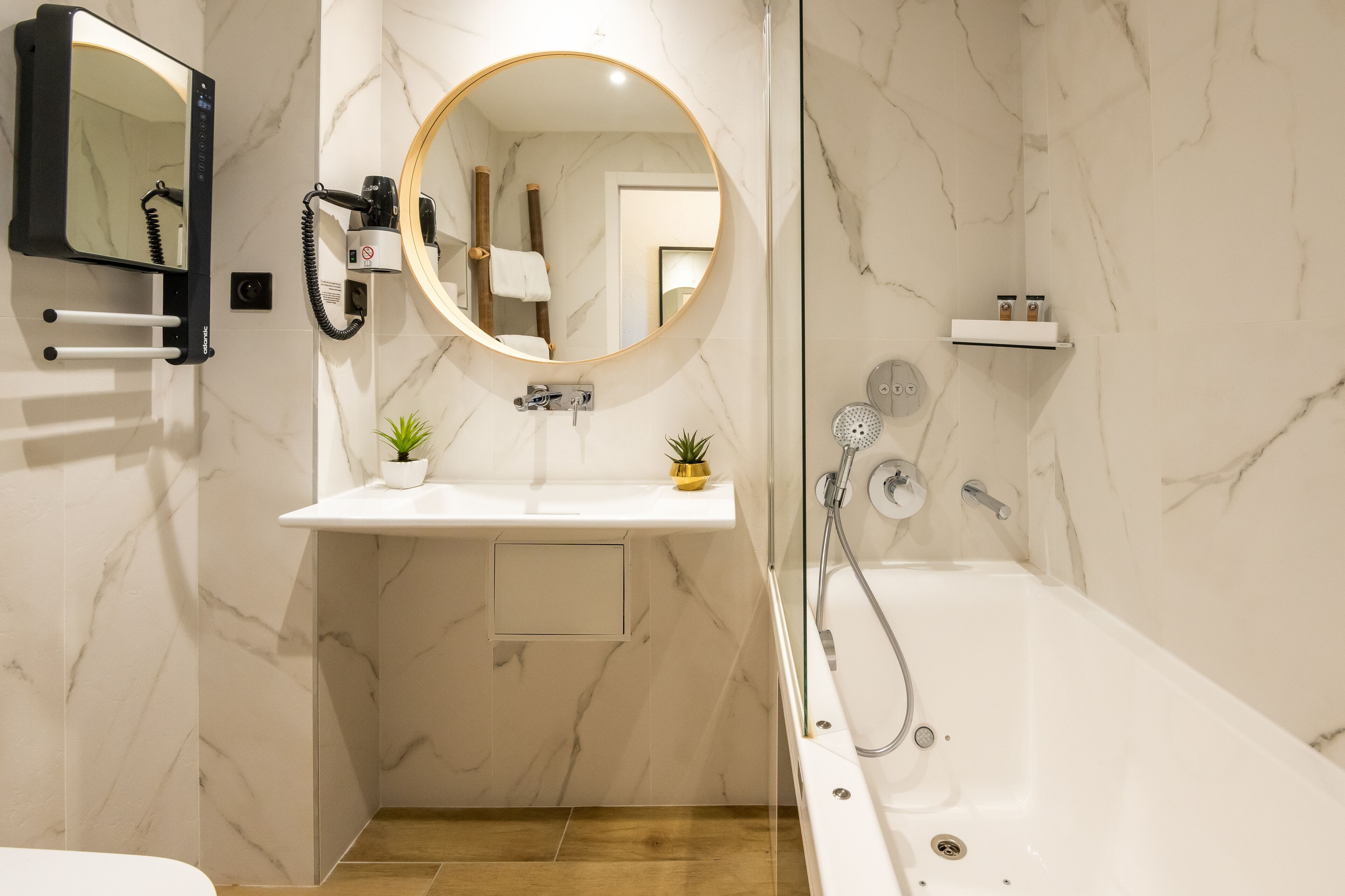 Un baño moderno con pared de mármol, un espejo redondo, un lavabo blanco, un secador de pelo de mano y una bañera.