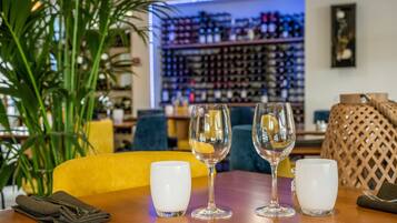 Una mesa con tres copas de vino y dos tazas blancas, una planta y un fondo con estantes llenos de botellas.