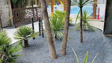 Un área exterior con jardines, palmeras, una piscina y una pérgola de madera.