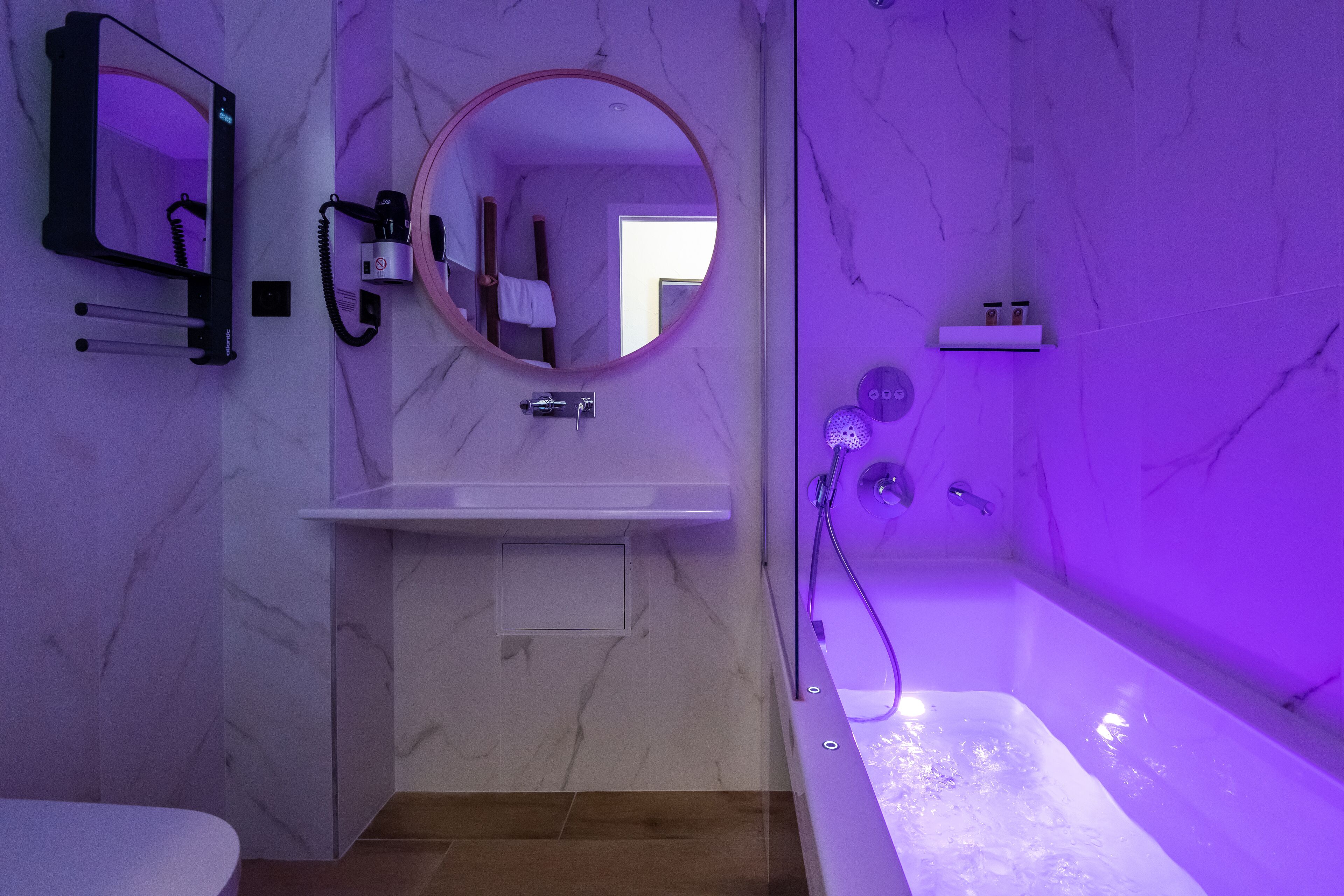 Bany amb paret de marbre, un mirall rodó, un lavabo, un vàter i una banyera amb il·luminació morada.