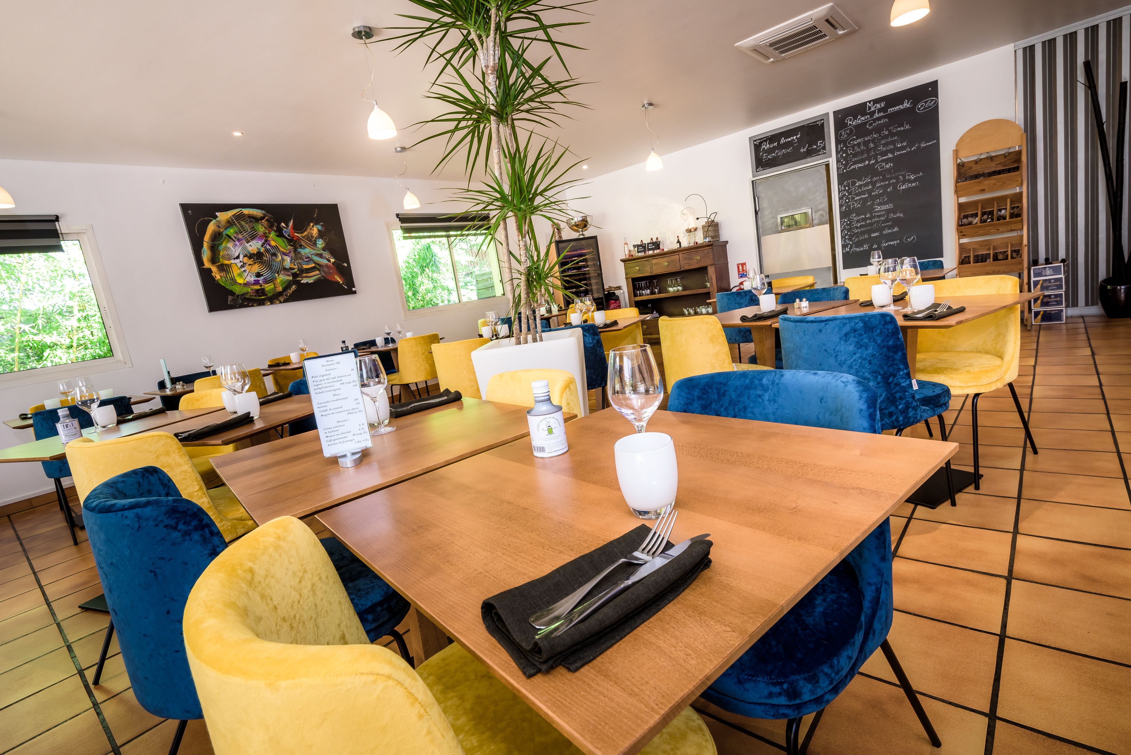 Un ristorante con sedie in velluto giallo e blu, tavoli di legno, una pianta grande e una lavagna con il menu.