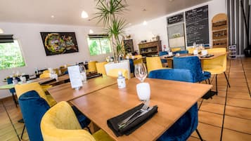 Un ristorante con sedie in velluto giallo e blu, tavoli di legno, una pianta grande e una lavagna con il menu.