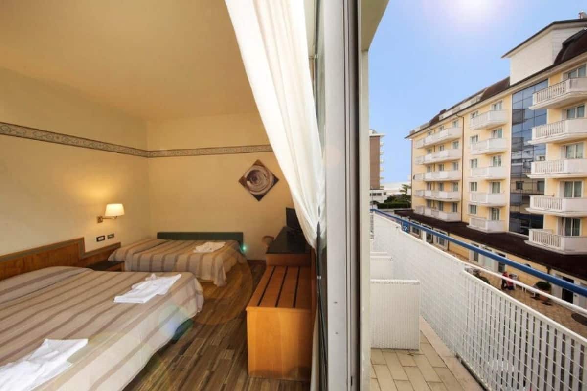 A hotel room with two beds, a wooden bench, a wall-mounted TV, and a balcony with a railing.
