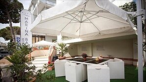 A covered outdoor seating area with white furniture and a large umbrella. - Hotel Marina Bay (Rimini)