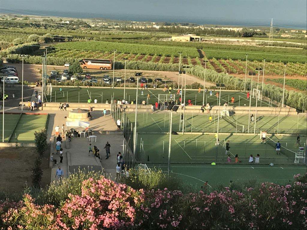 A sports field with people playing soccer, surrounded by green fields and a parking area with vehicles.