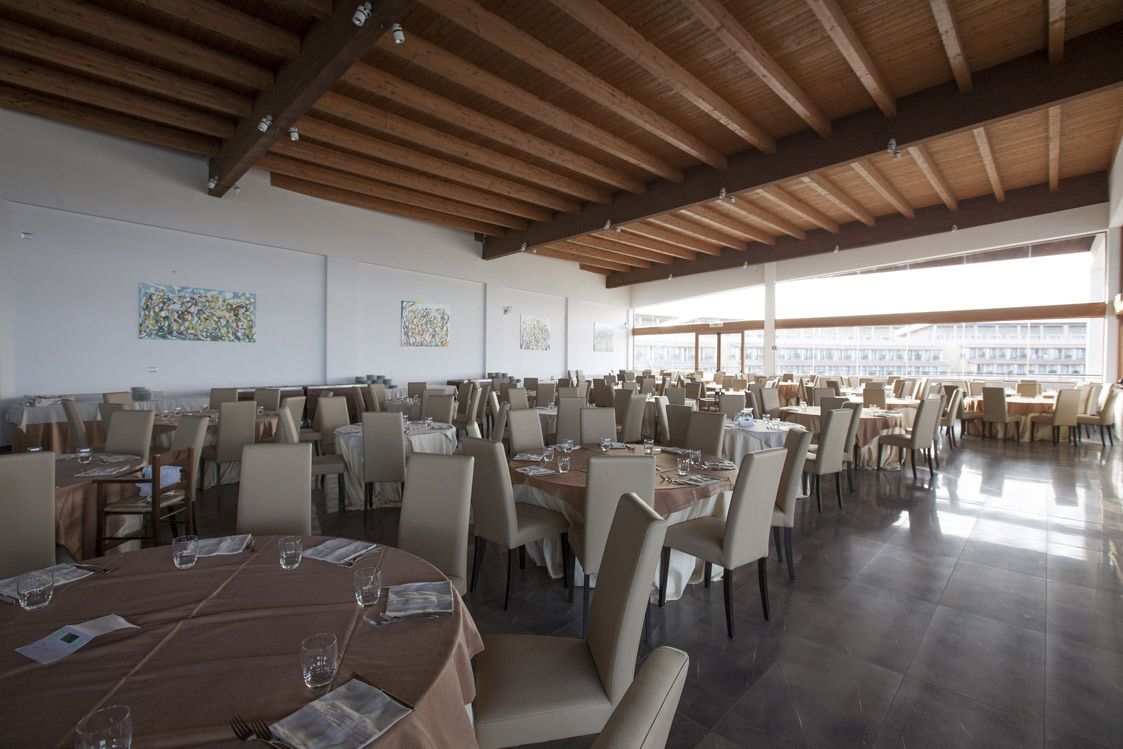 A spacious dining area with multiple tables and chairs, wooden ceiling beams, and large windows allowing natural light.