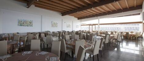 A spacious dining area with multiple tables and chairs, wooden ceiling beams, and large windows allowing natural light.