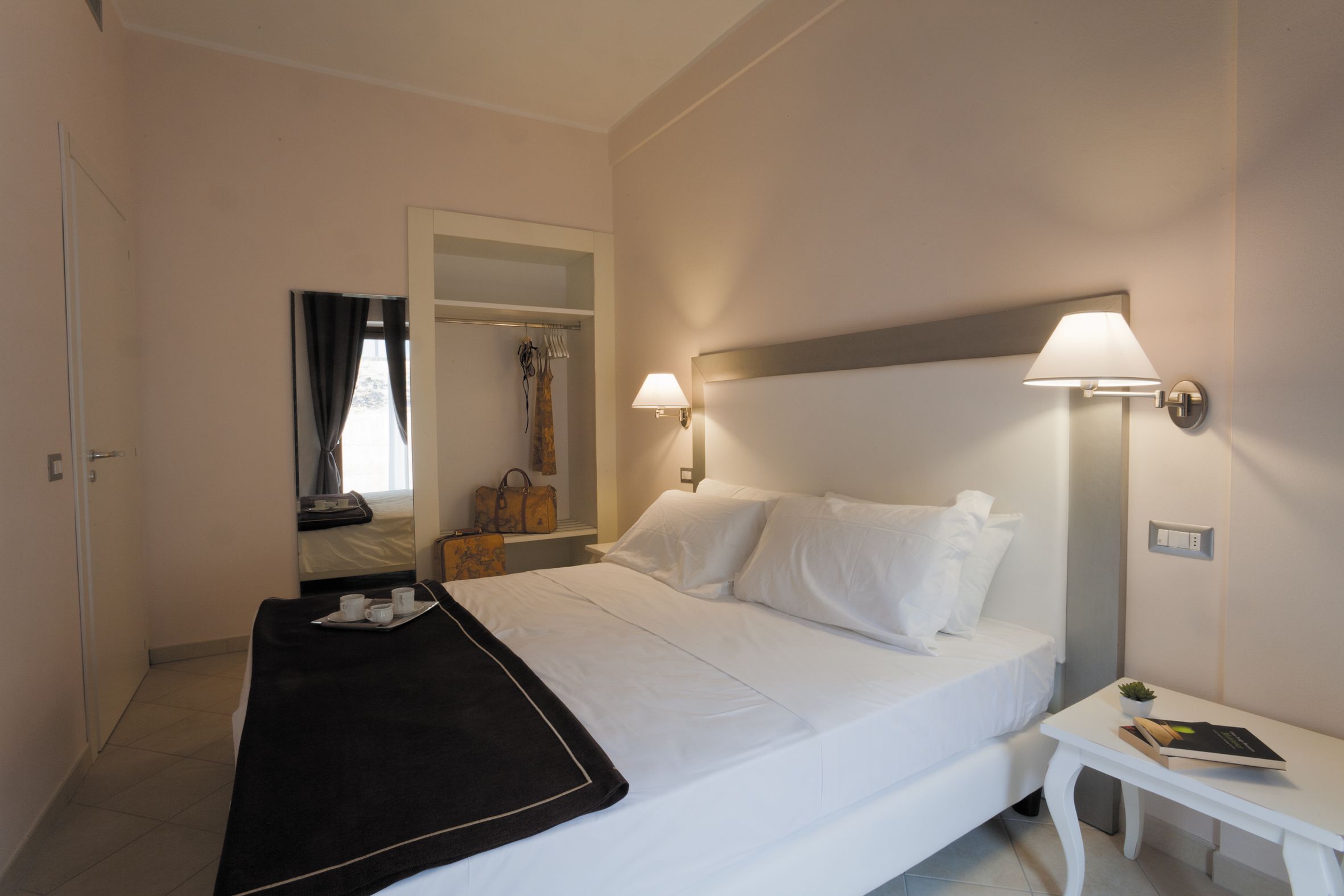 Camera d'albergo con un letto, un comodino, una lampada e vista su un armadio con uno specchio e una borsa.