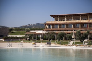 A pool area with lounge chairs and umbrellas, a building with multiple balconies, and a view of hills in the background.