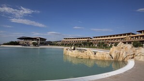 Un hotel con una piscina amplia, una fuente rocosa y un edificio moderno con varios balcones.
