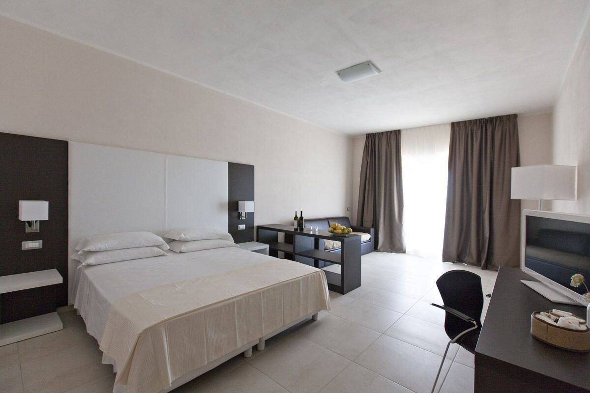 A modern hotel room with a large bed, a desk, a chair, a TV, and a small table with a lamp.