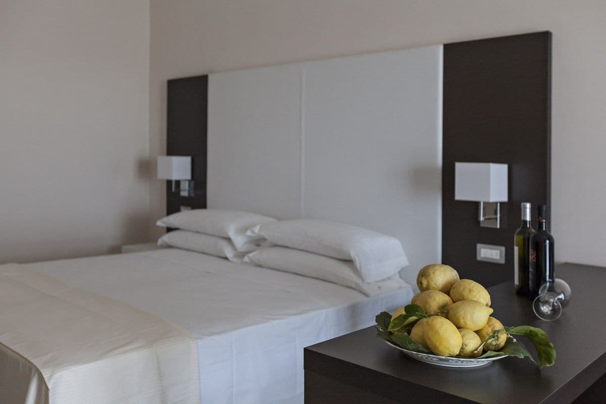 A hotel room with a neatly made bed, a bedside table with a bowl of lemons and a bottle of olive oil, and a wall-mounted lamp.