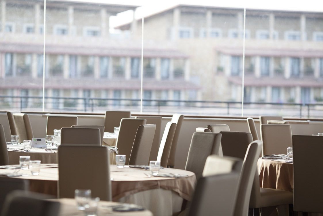 Een restaurant met ronde tafels en stoelen, grote ramen met uitzicht op gebouwen.