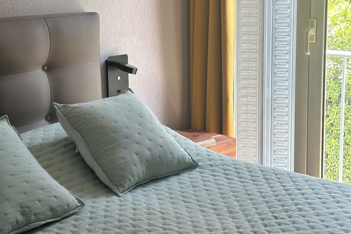A bedroom with a large bed, two pillows, a headboard, a window with curtains, and a thermostat on the wall.