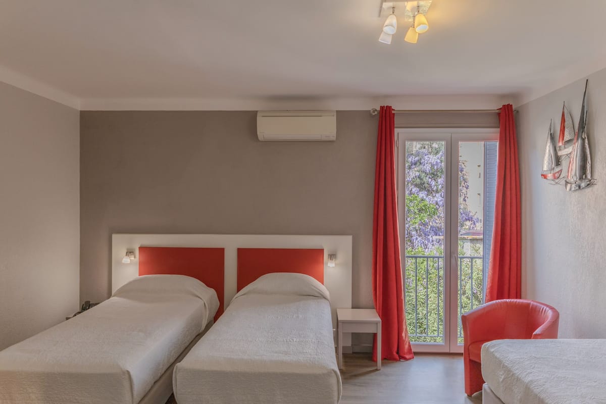 A hotel room with two single beds, a small table, a chair, and a balcony with red curtains.
