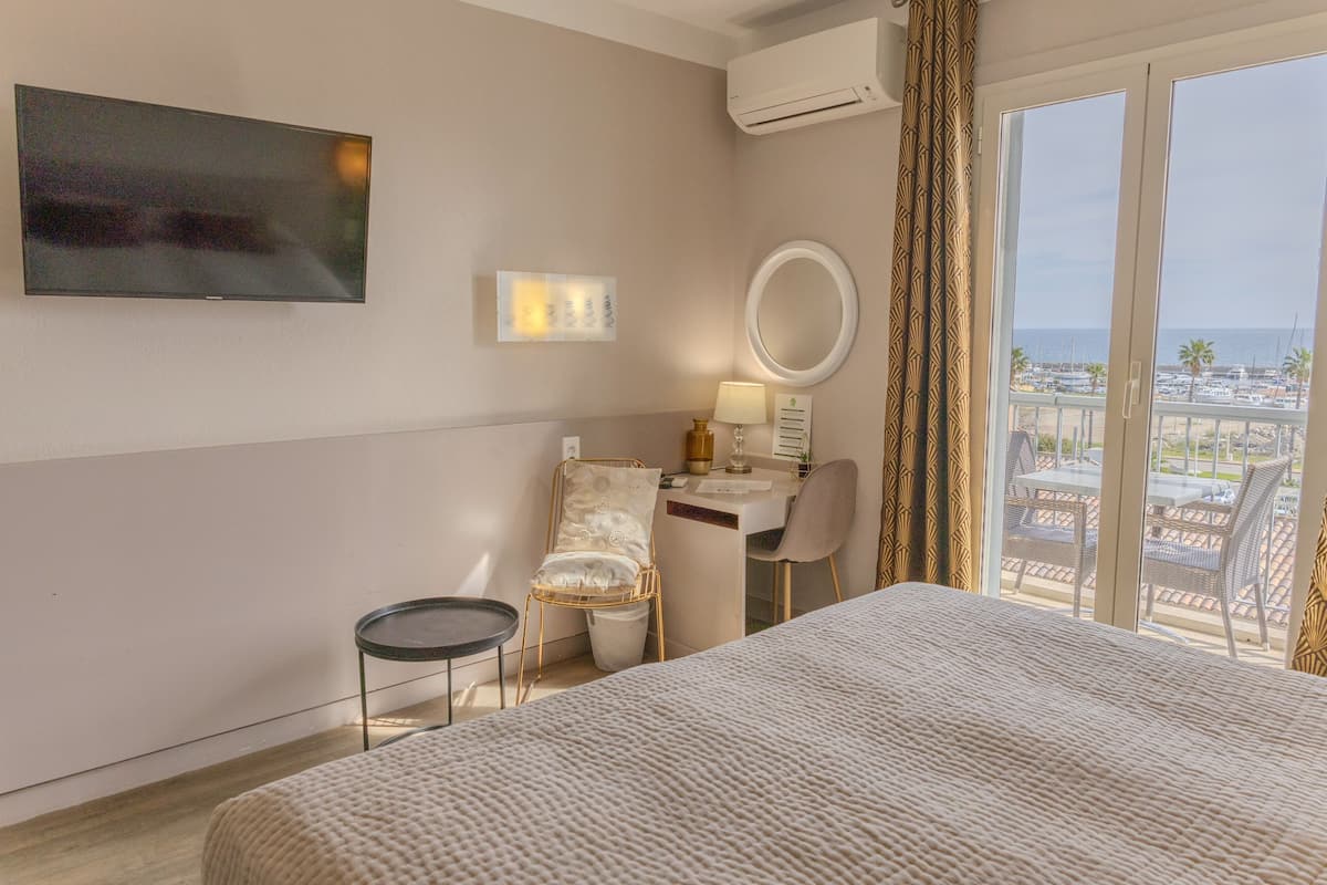 A hotel room with a bed, a desk with a chair, a TV, a window with a view of the beach and boats, and an air conditioning unit.