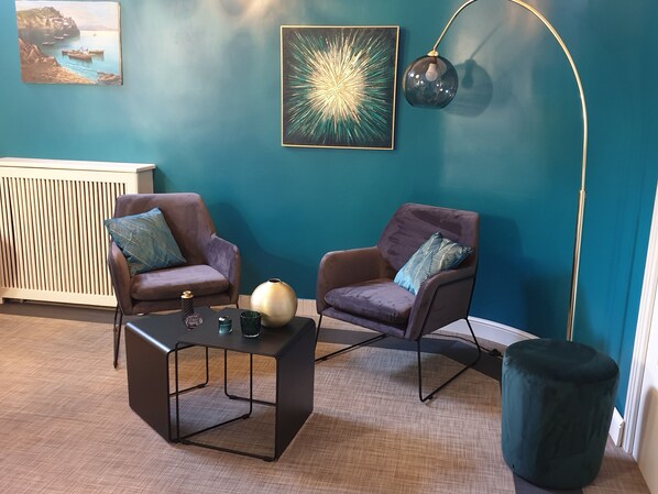Ein Zimmer mit türkisfarbenen Wänden, zwei braune Sessel mit gemusterten Kissen, ein schwarzer Couchtisch, eine Stehlampe und eine Heizung.