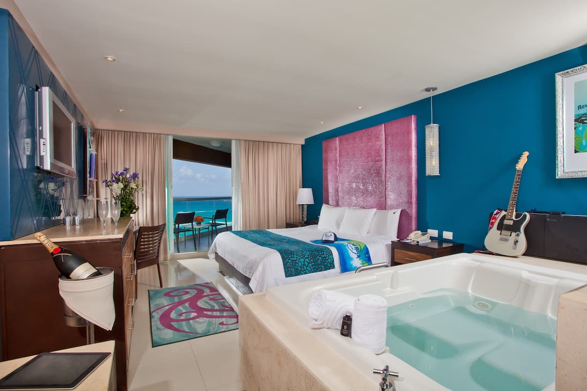 A hotel room with a bed, a bathtub, a desk with a computer, a guitar, and a view of the ocean.