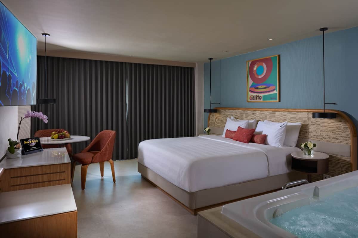 A hotel room with a bed, a desk, a chair, a small table with fruit, a wall-mounted TV, a painting, and a bathtub.
