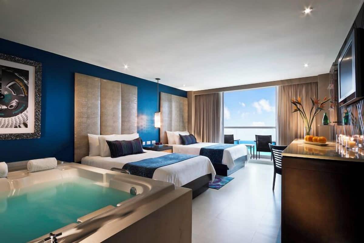 A hotel room with two beds, a large window, a bathtub, and a bar area.