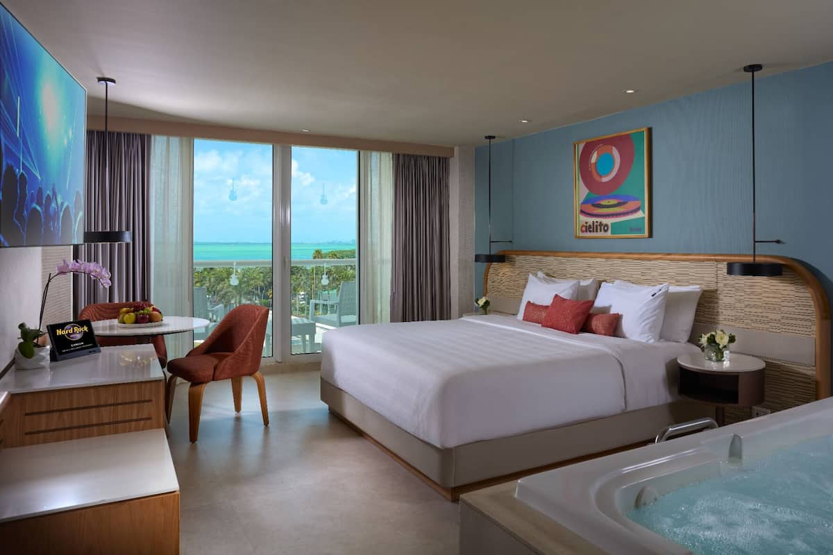 A hotel room with a large bed, a sitting area with a chair and table, a small round table with a vase, a large window with a view of the sea, and a jacuzzi tub.