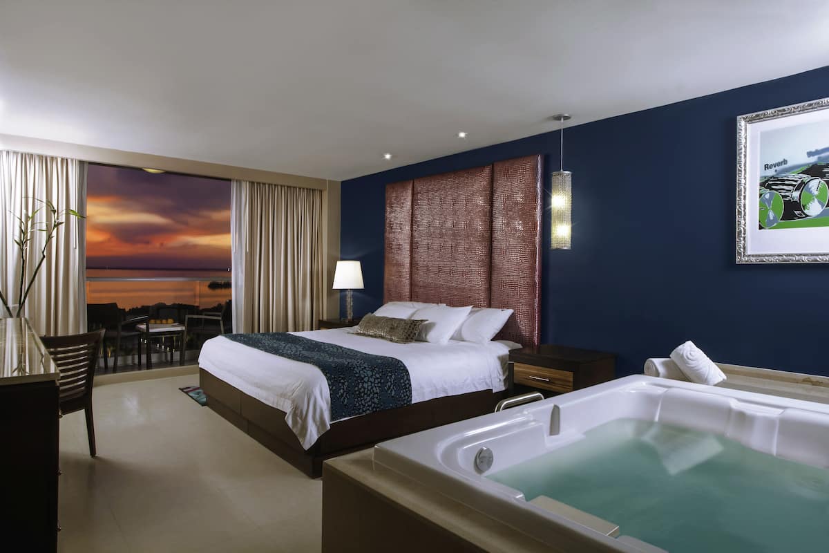 A hotel room with a bed, a desk, a chair, a large window with curtains, a view of the sunset, a wall-mounted artwork, and a bathtub.