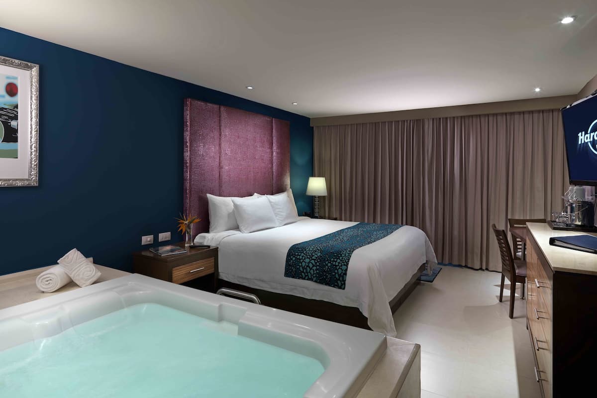 A hotel room with a large bed, a desk, a chair, a desk lamp, a TV, a bathtub, and a framed picture on the wall.