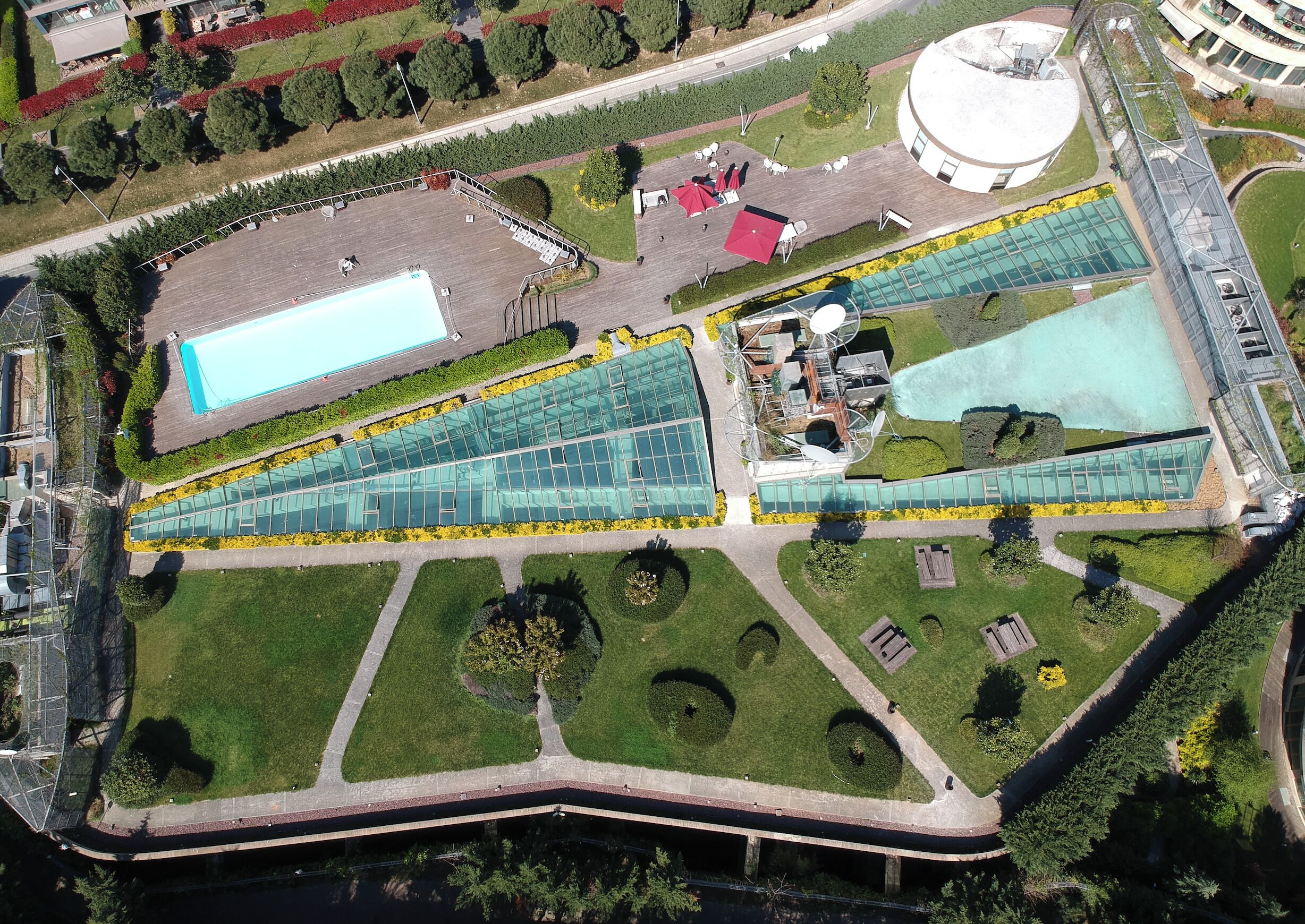 Vista aérea de un edificio moderno con una amplia fachada de vidrio, piscina y zonas ajardinadas alrededor.