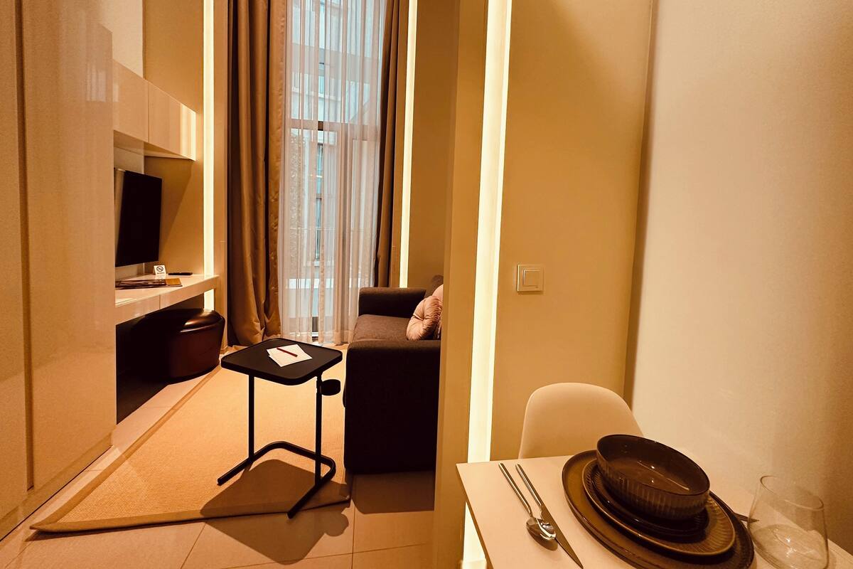 Habitación de hotel compacta con cama, un pequeño escritorio con silla, televisión, ventana con cortinas y zona de comedor con mesa para dos.