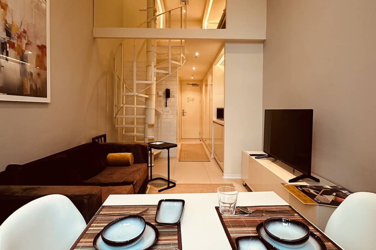 Una sala de estar moderna con un sofá, una mesa de comedor preparada para dos personas, un televisor y una escalera que sube a un nivel superior.