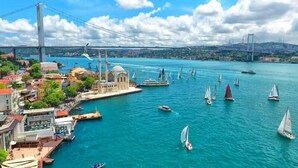 A coastal cityscape with a prominent mosque, a bridge, and a marina with sailboats. - Ortaköy Aparts (Istanbul)