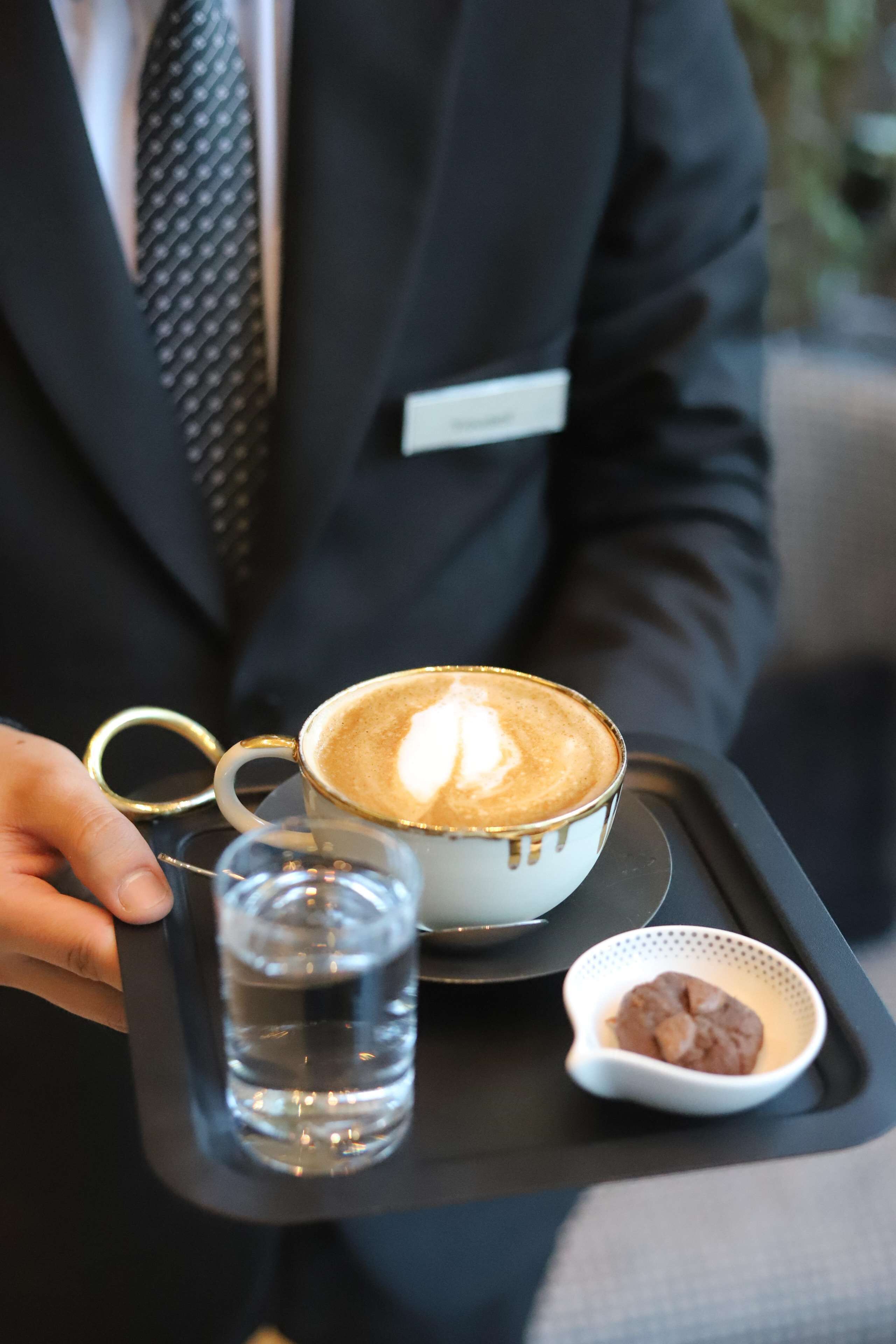 A person holding a tray with a cup of coffee, a glass of water, and a small bowl of chocolate.