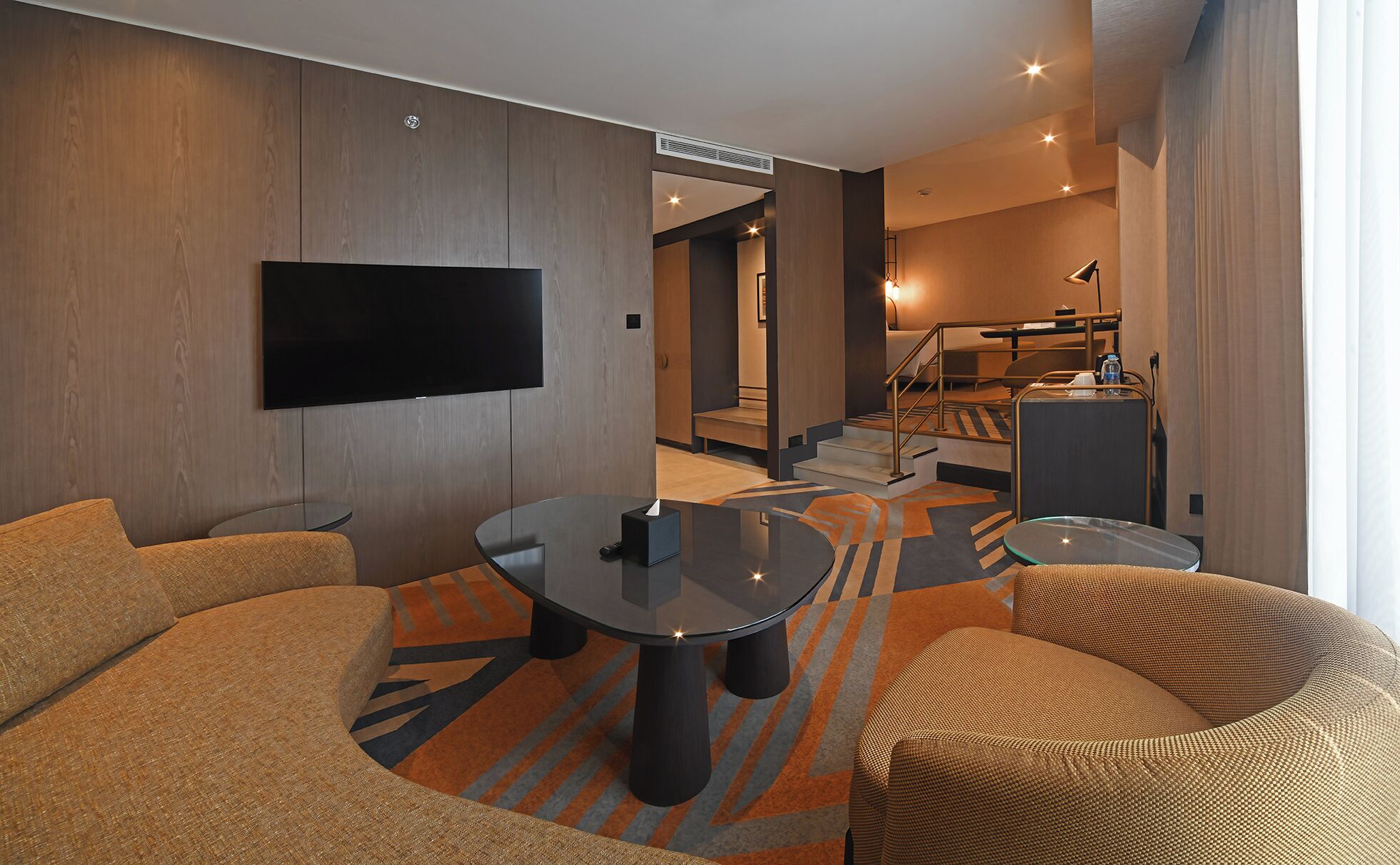 A modern hotel room with a flat-screen TV, a round glass table, and a sofa. There is a staircase leading to an upper level.