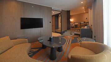 A modern hotel room with a flat-screen TV, a round glass table, and a sofa. There is a staircase leading to an upper level.
