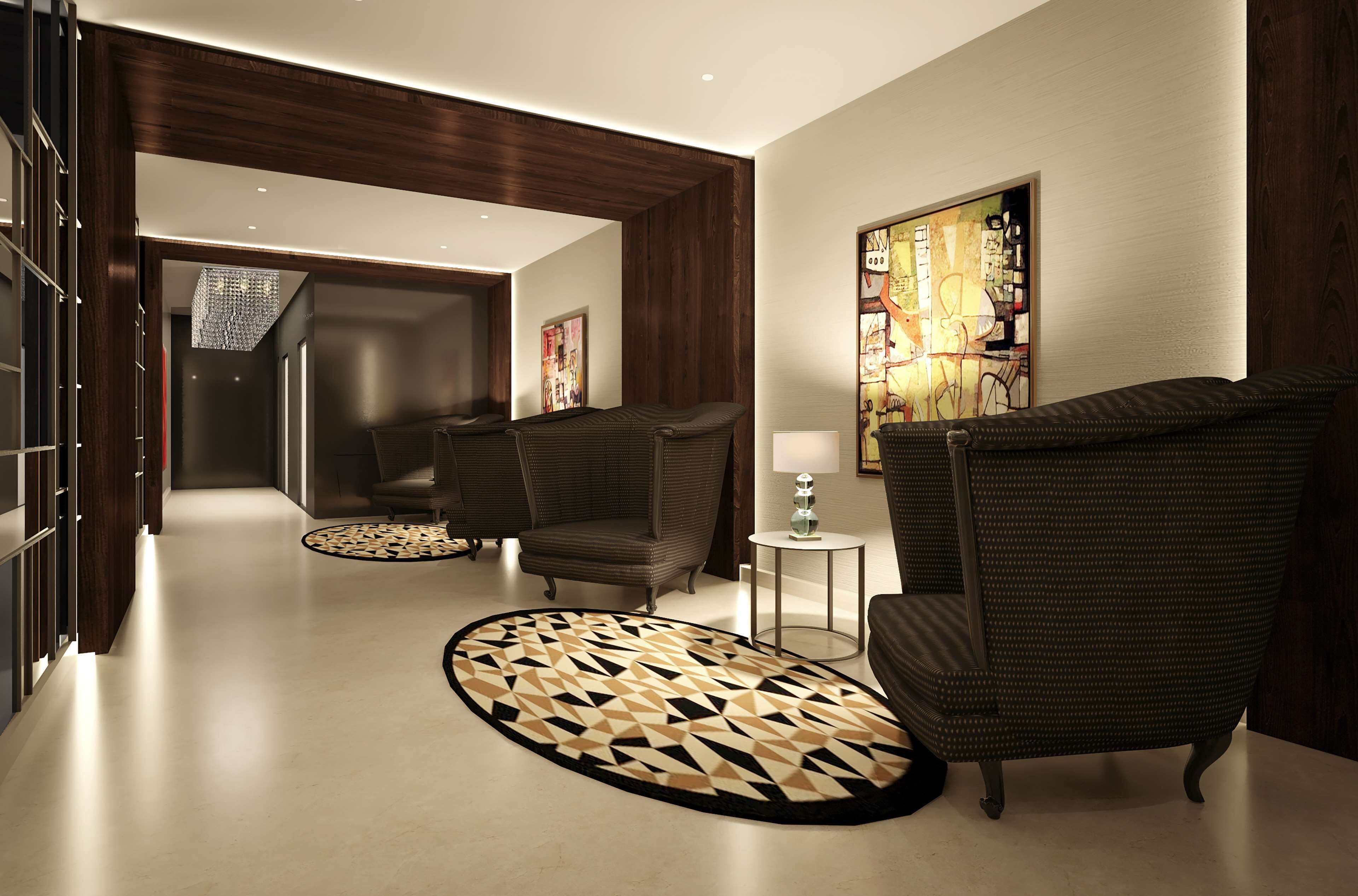 A modern hotel lobby with a geometric patterned rug, dark upholstered chairs, a small side table with a lamp, and a large abstract painting on the wall.
