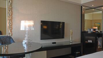 A modern hotel room with a flat-screen TV mounted on the wall, a glass table with a decorative lamp, and a minibar.
