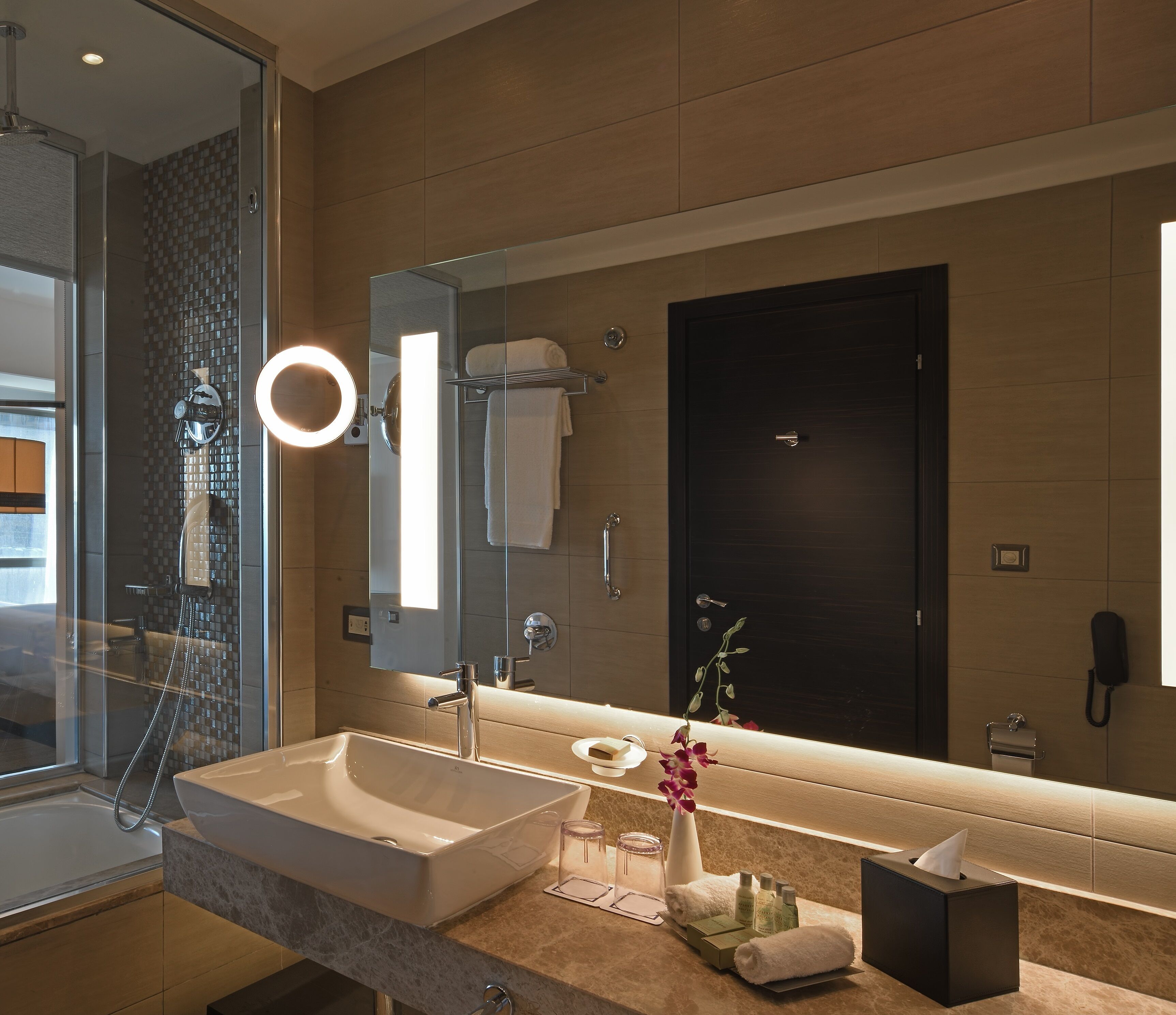 A modern bathroom with a large mirror, a glass shower enclosure, a white sink, and a vanity with toiletries.