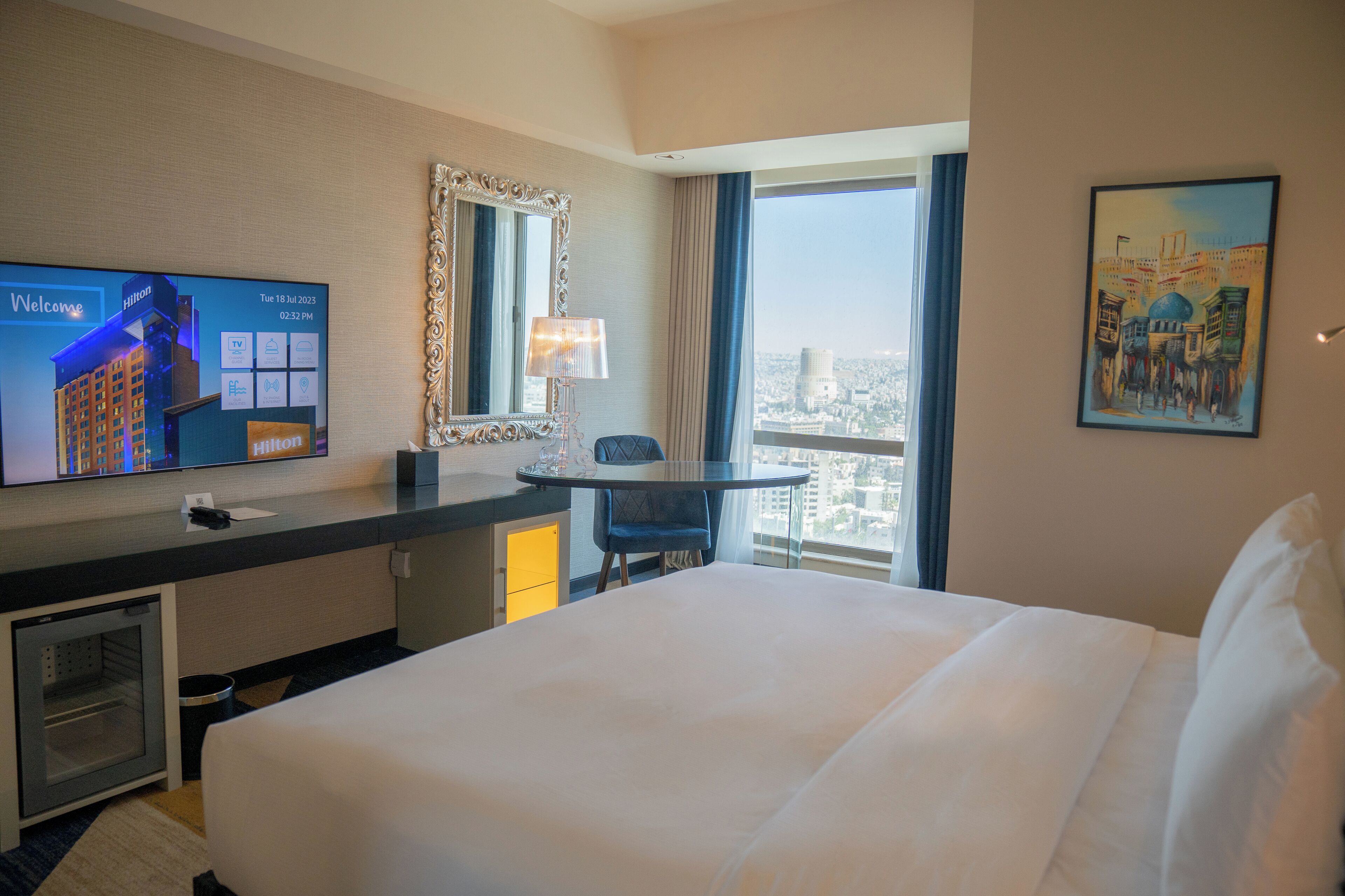 Habitación de hotel con cama, escritorio con televisor mostrando el logotipo de Hilton, vista a la ciudad, un cuadro en la pared y ventana con cortinas.