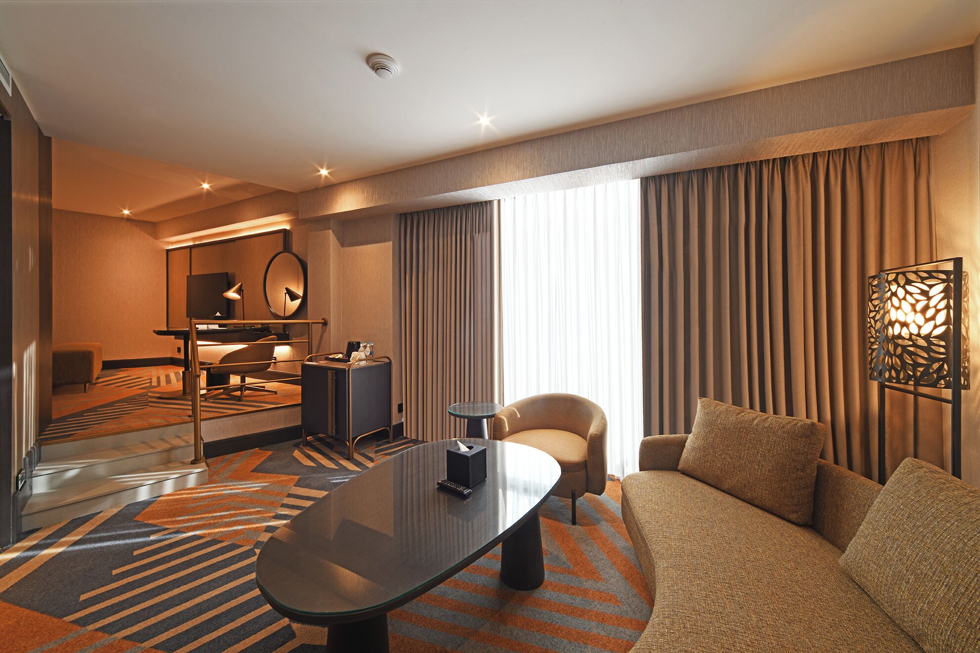 A modern hotel room with a sofa, armchair, and a glass-top table. There is a desk with a chair, a mirror, and a lamp. The room has a large window with curtains and a decorative wall panel.