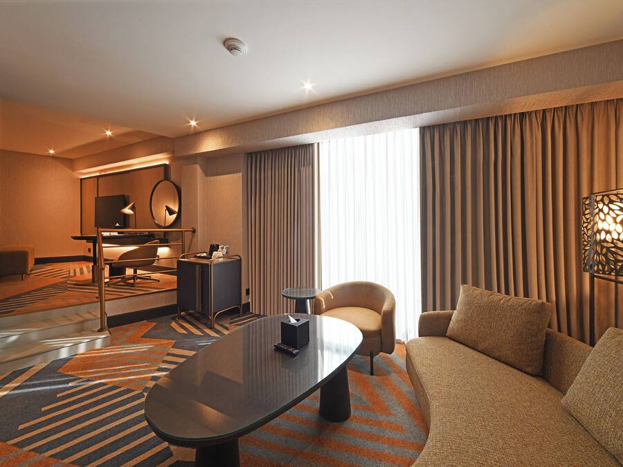 Een moderne hotelkamer met een bank, fauteuil en een tafel met glazen blad. Er is een bureau met stoel, een spiegel en een lamp. De kamer heeft een groot raam met gordijnen en een decoratief wandpaneel.