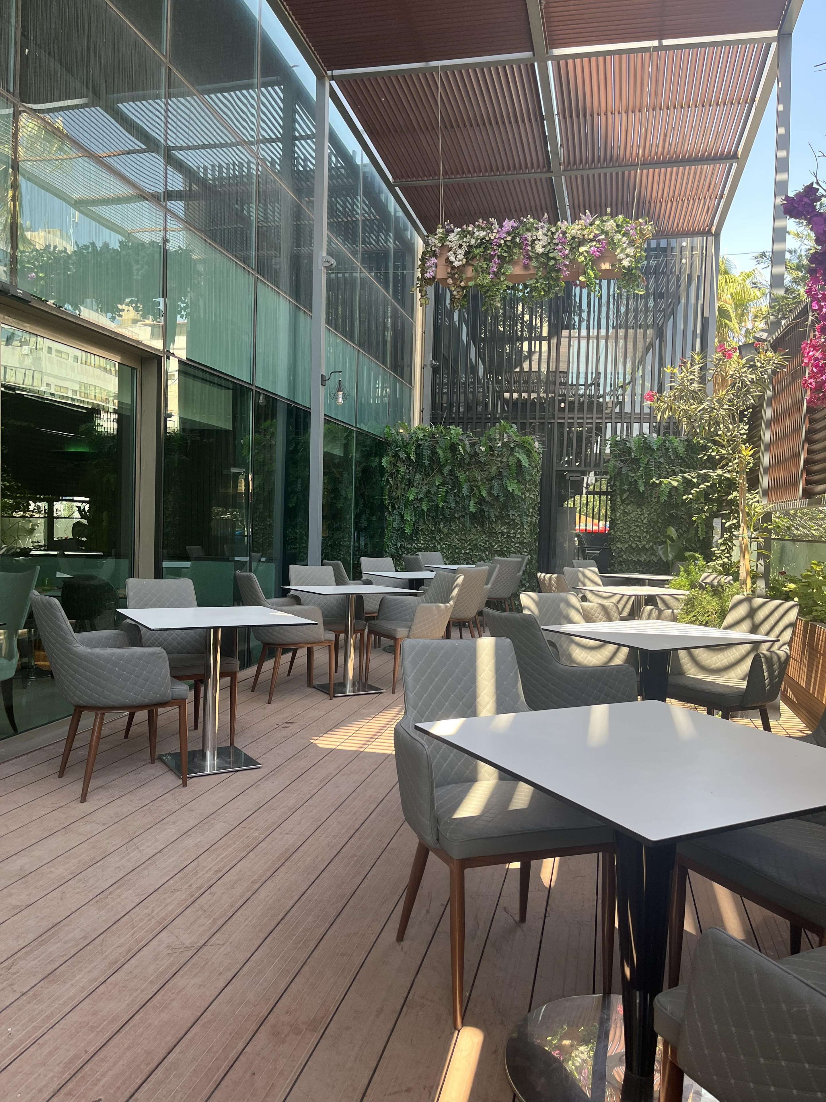 Un patio al aire libre con mesas y sillas, una terraza de madera y un muro de vidrio que refleja el entorno.