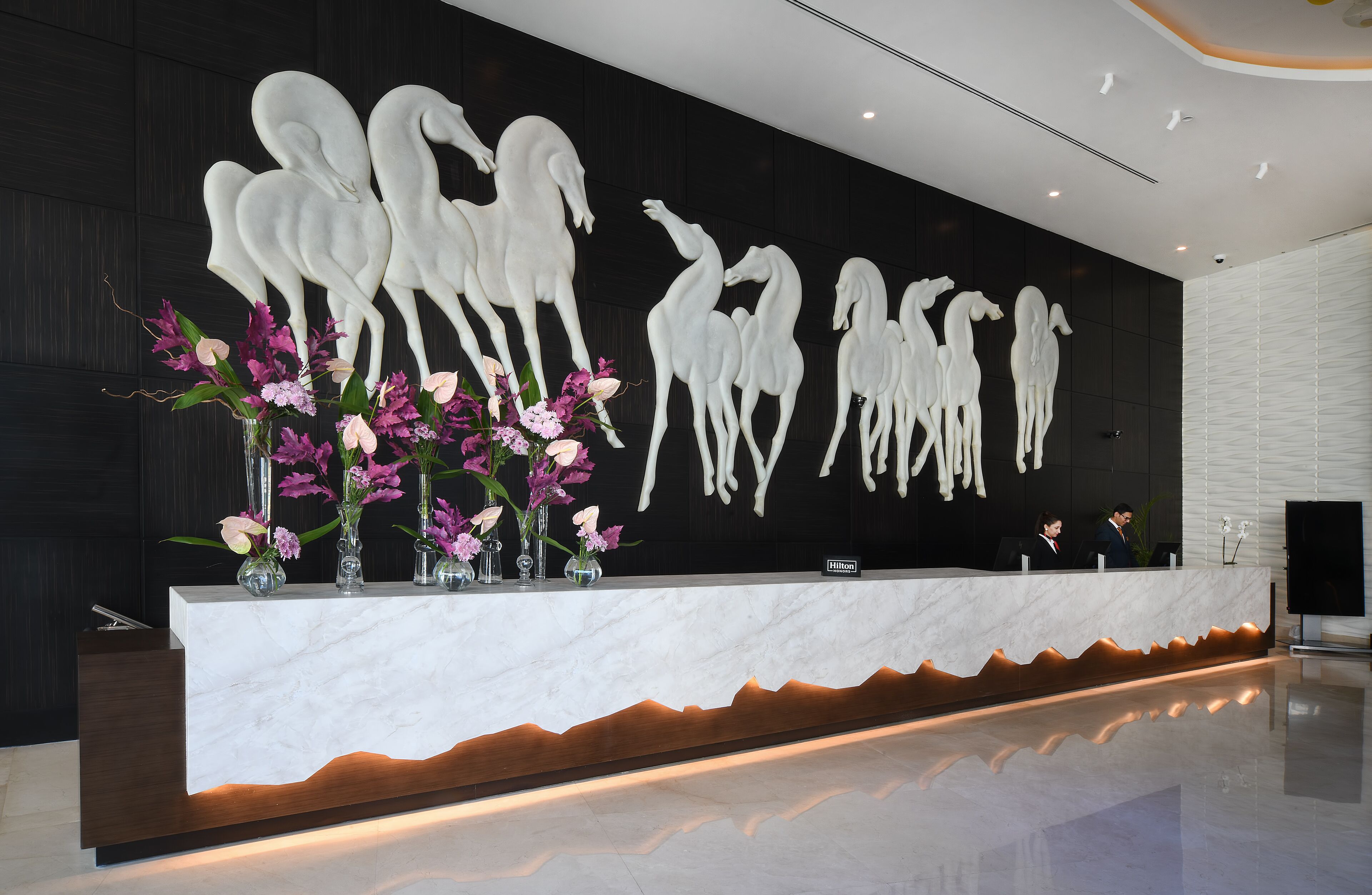 Un comptoir d’accueil d’hôtel avec un plateau en marbre, un mur noir orné de silhouettes de chevaux blancs et une composition florale.