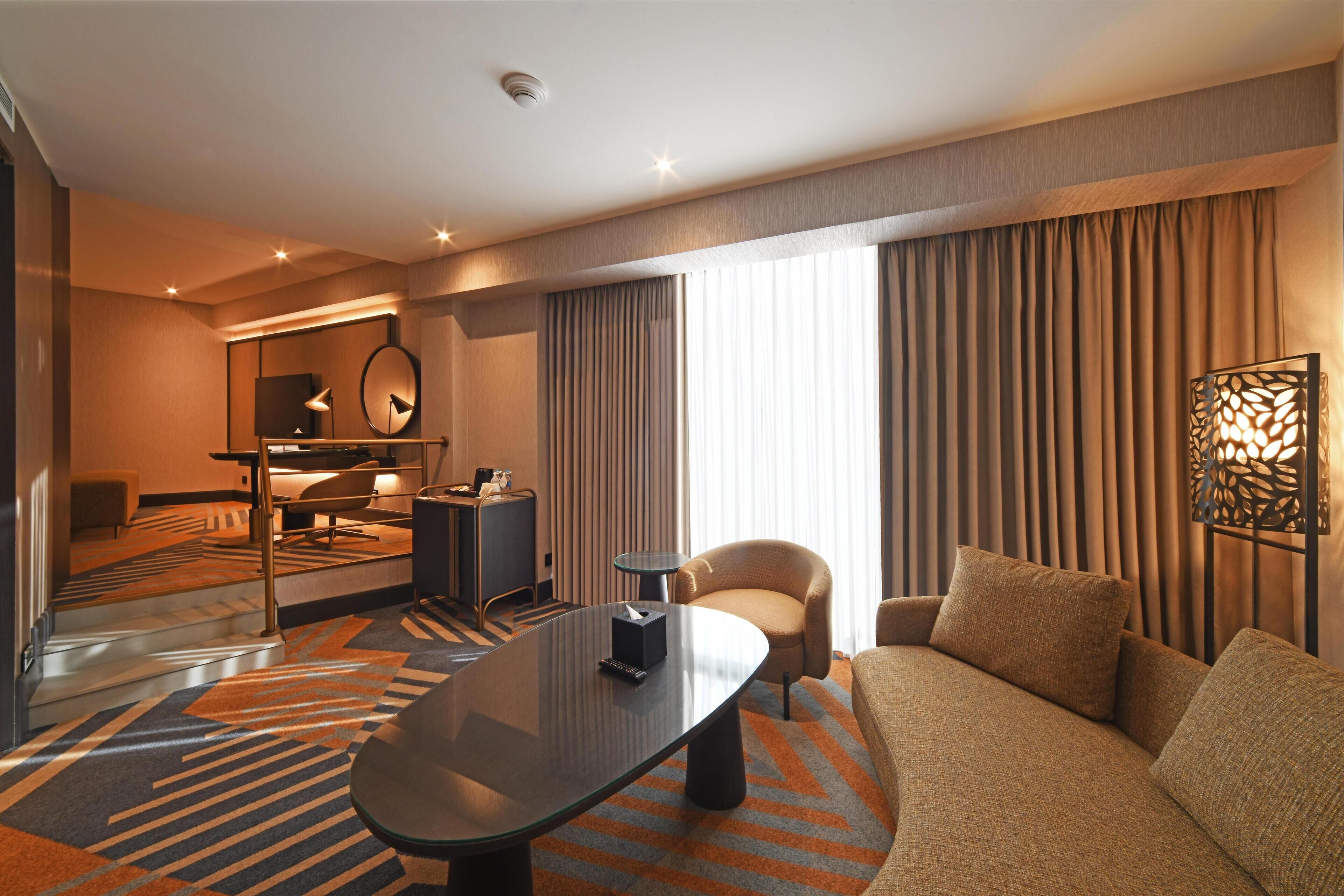 Una habitación de hotel moderna con sofá, una mesa de centro de vidrio, un escritorio con silla, un espejo y un ventanal con cortinas.