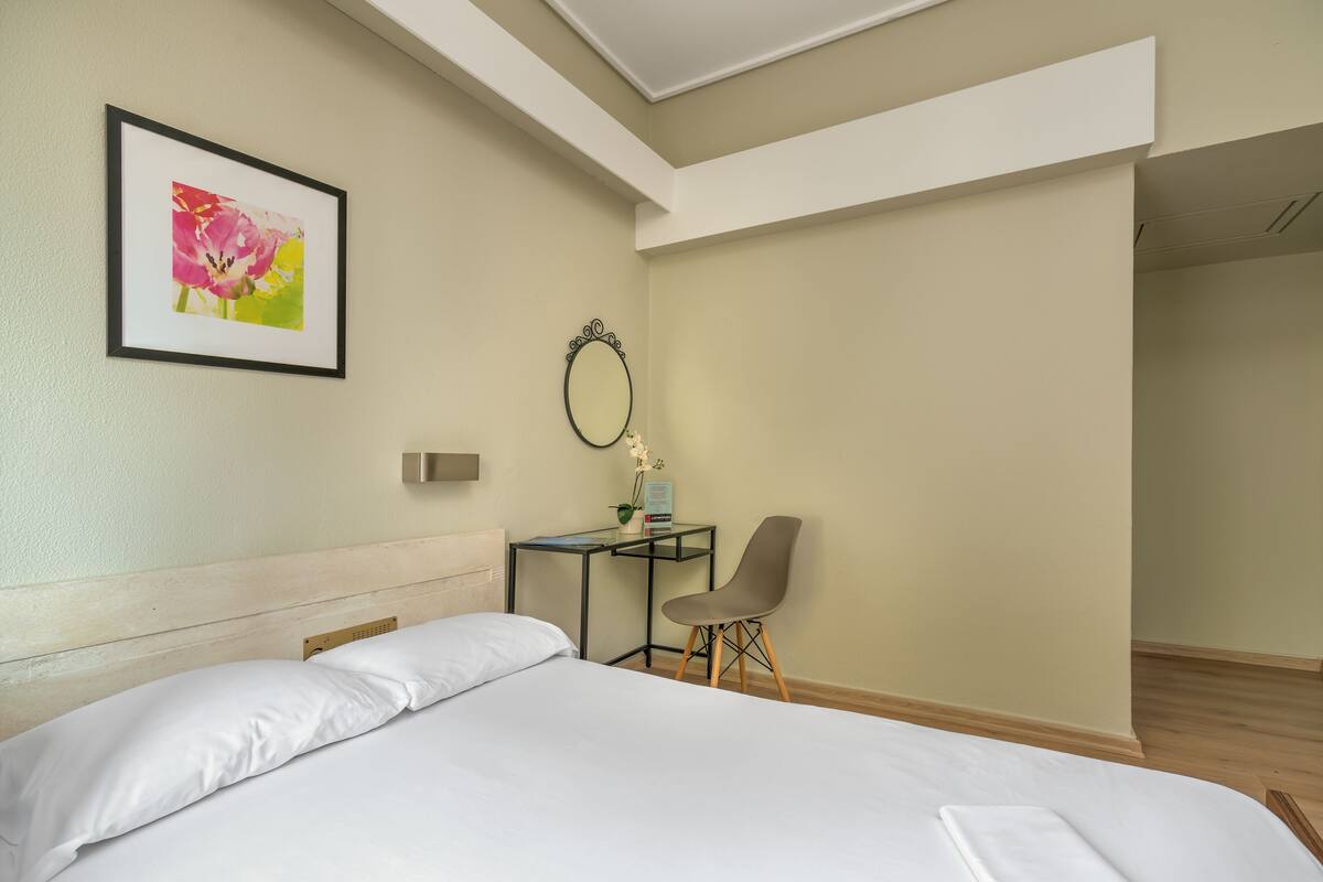 A hotel room with a bed, a desk, a chair, a framed picture on the wall, and a mirror.