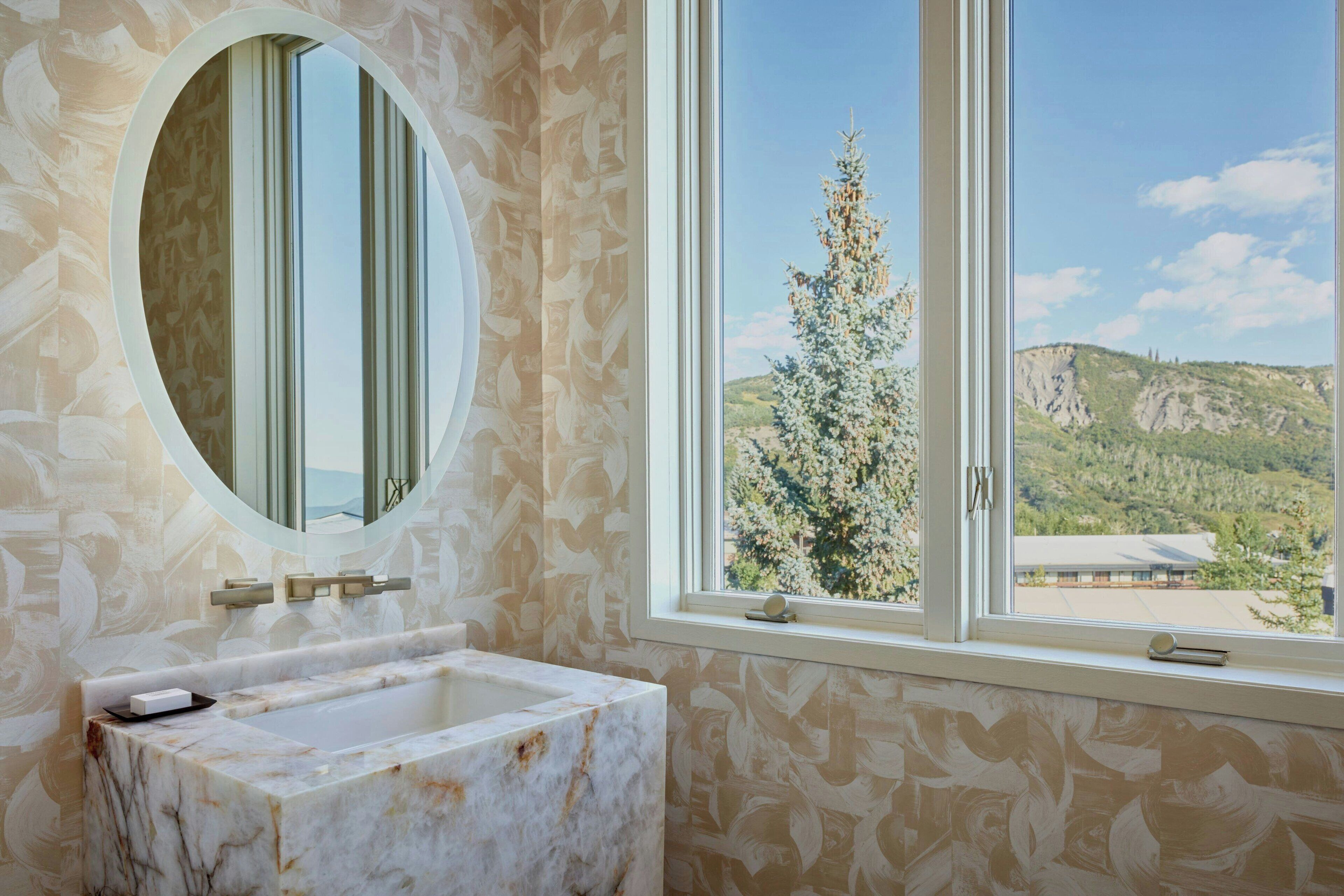 Une salle de bain avec un lavabo en marbre, un miroir rond et une fenêtre donnant sur un arbre et des montagnes.