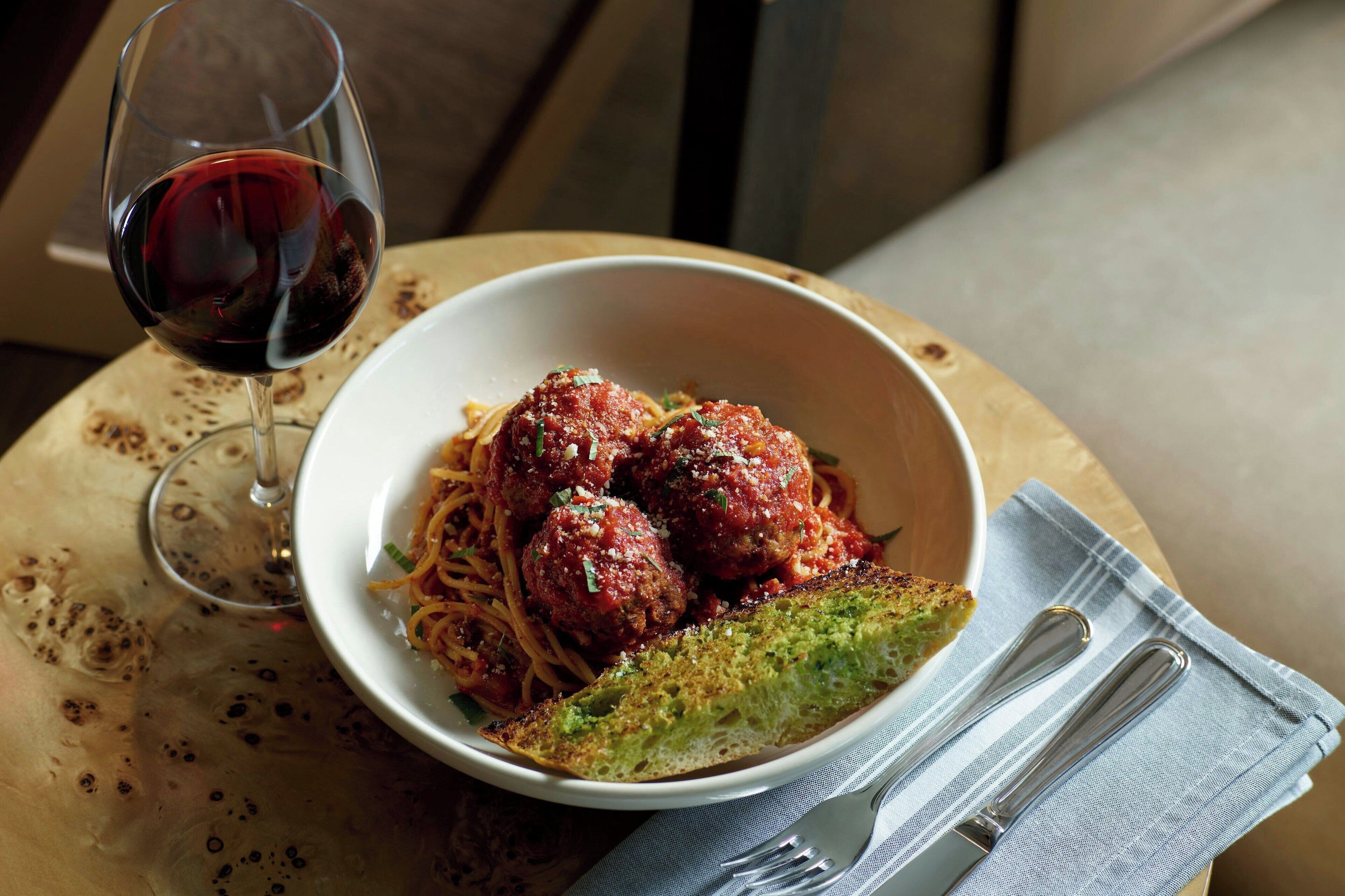 A bowl of spaghetti with meatballs and a glass of red wine on a wooden table.